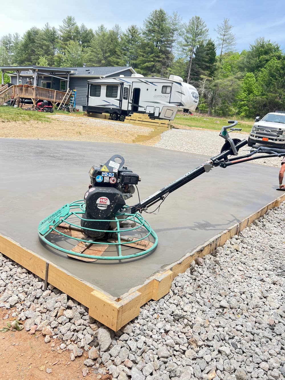 Concrete finishing machine on newly poured slab with RV and wooded area in the background.