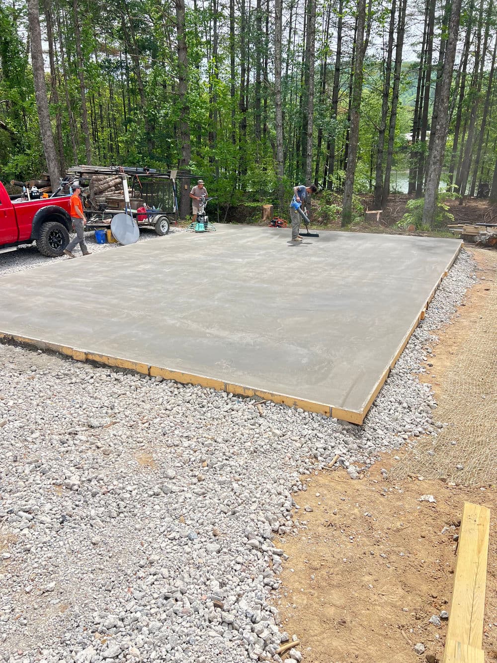 Concrete slab being poured with workers and equipment in a wooded construction site.