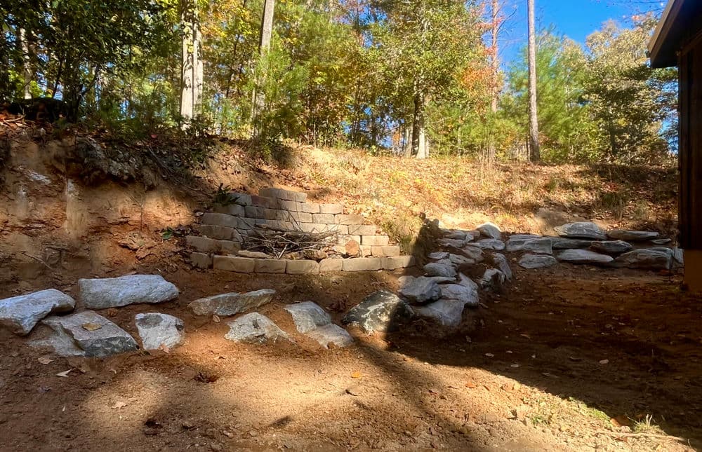 Stone fire pit surrounded by rocks in a wooded outdoor landscape with autumn foliage.