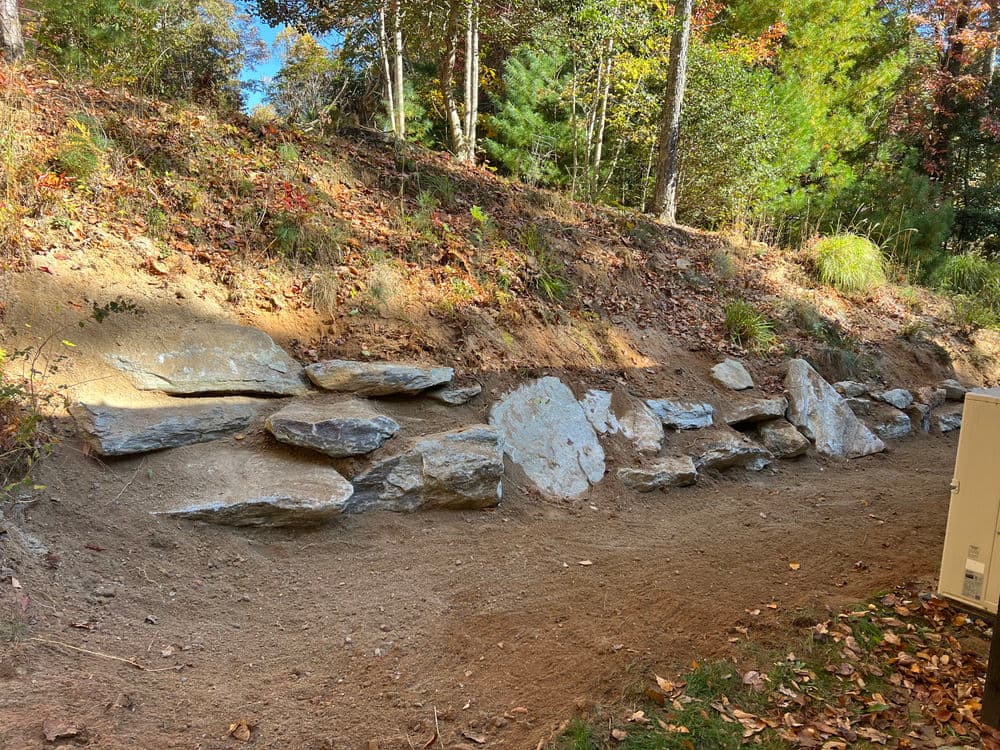 Natural stone retaining wall along a dirt path in a wooded area with autumn foliage.