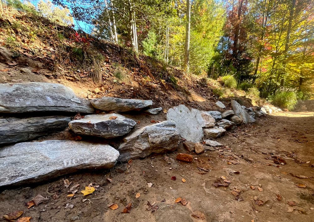 Rock retaining wall along a dirt path surrounded by autumn foliage and trees.