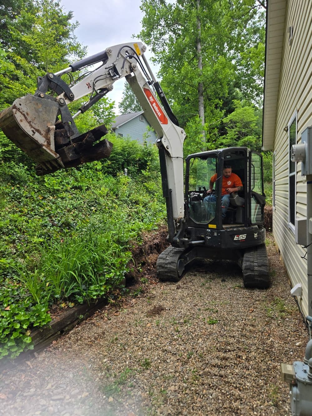Excavator digging near a house with greenery in the background on a gravel pathway.