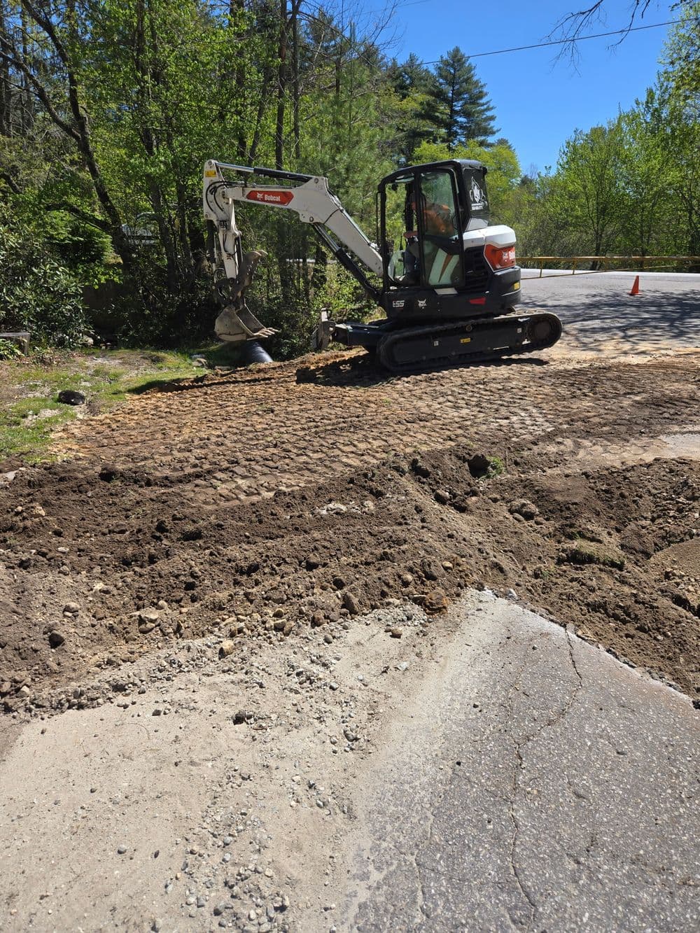 Excavator working on a road repair project surrounded by trees and freshly dug soil.