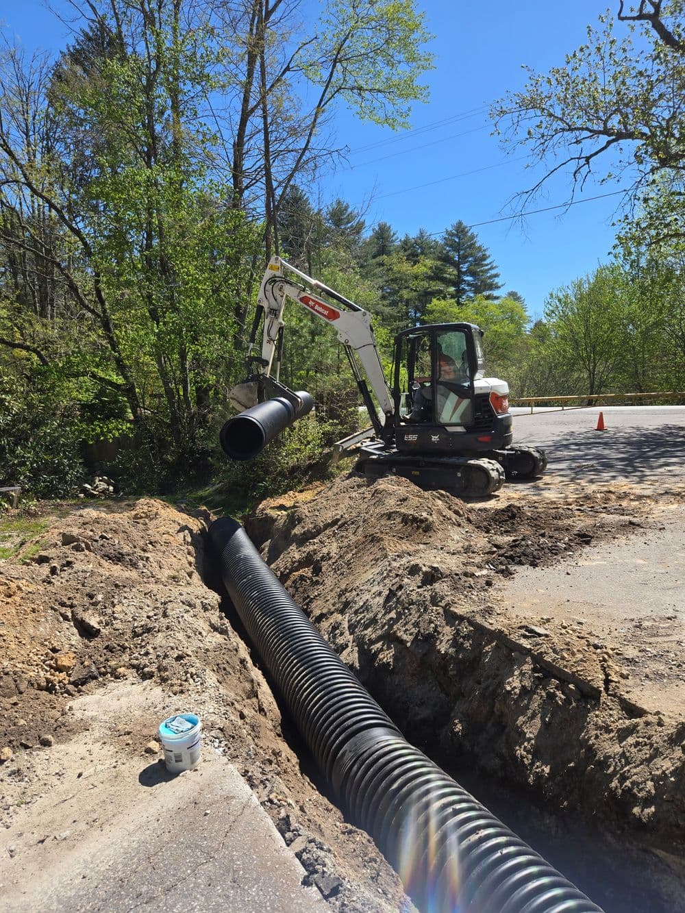 Excavator installing black drainage pipe near roadside in a wooded area.