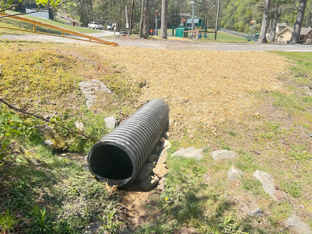 Drainage pipe in a forested area with playground equipment in the background.