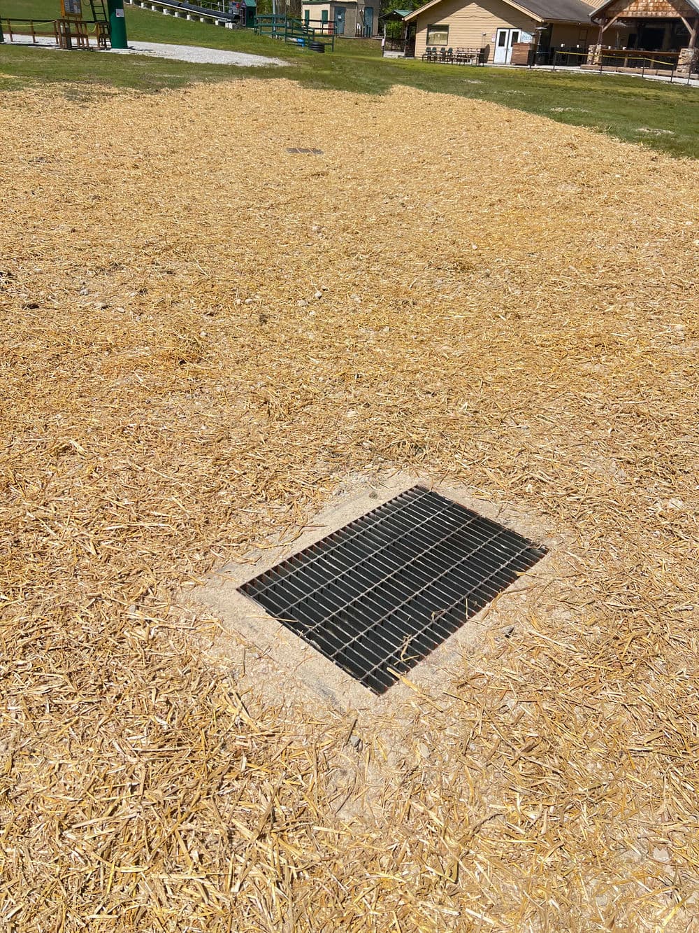 Grated drain cover on a sandy playground with straw scattered around.
