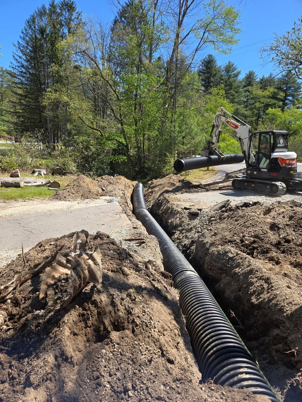 Excavator installing drainage pipe in a trench surrounded by trees and earthworks.