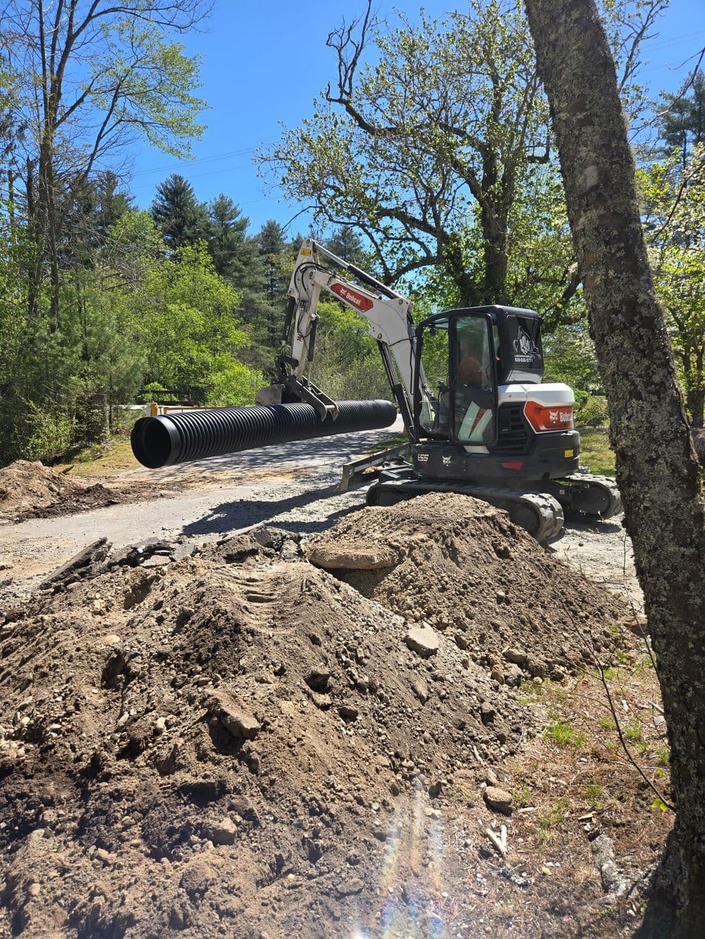 Excavator installing large drainage pipe on a dirt road surrounded by trees.