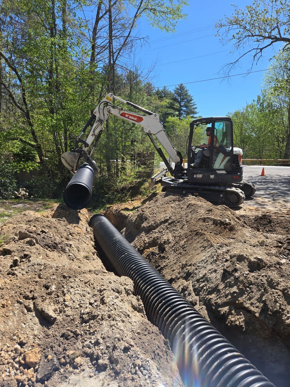 Excavator installing black drainage pipe in a trench surrounded by trees and blue sky.