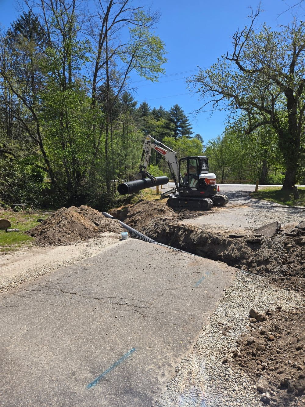 Excavator installing a pipe on a rural road surrounded by trees on a sunny day.