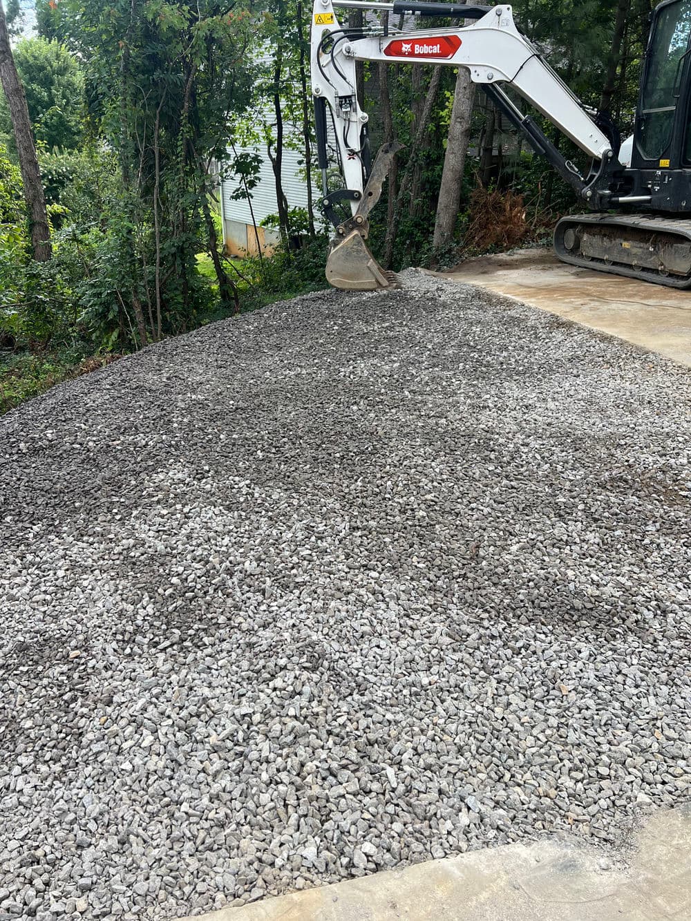 Bobcat machine pouring gravel on a prepared driveway surface surrounded by greenery.