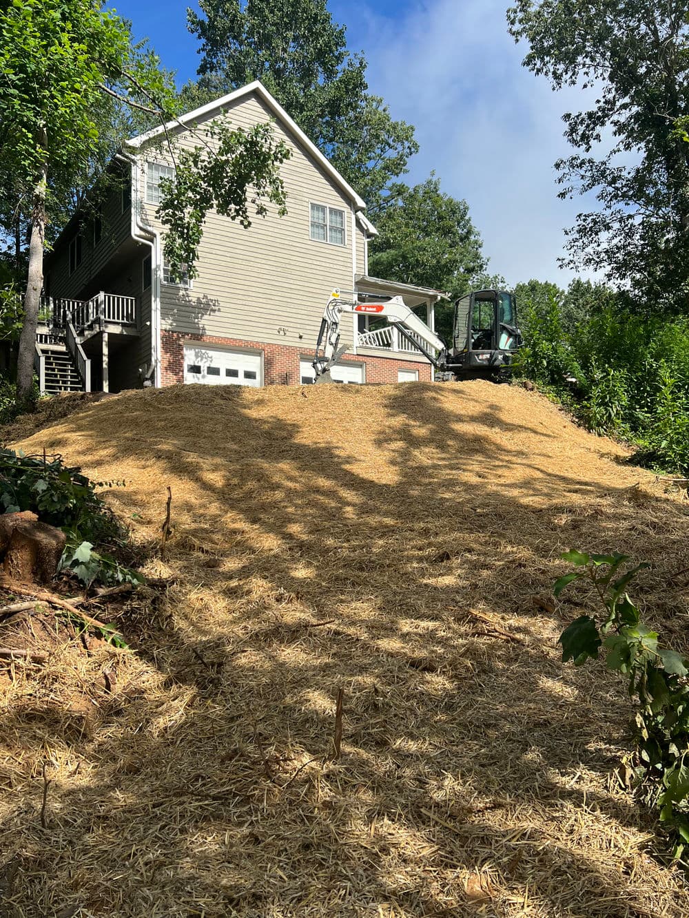 Home landscape with freshly laid straw mulch and equipment, surrounded by trees and blue sky.