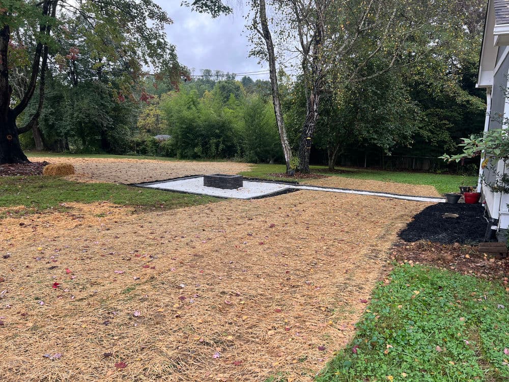 Backyard landscape with gravel fire pit area, surrounded by straw mulch and greenery.