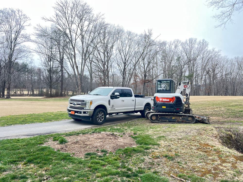 White Ford truck parked beside a compact excavator on a grassy area with bare trees.