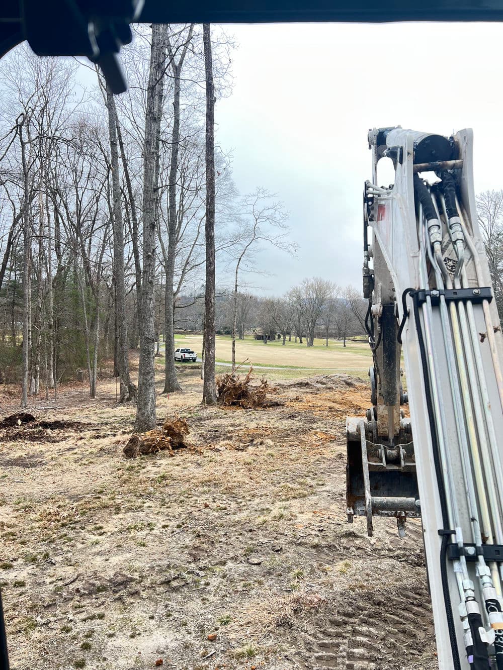 Excavator operator's view of cleared land with tree stumps and distant golf course.