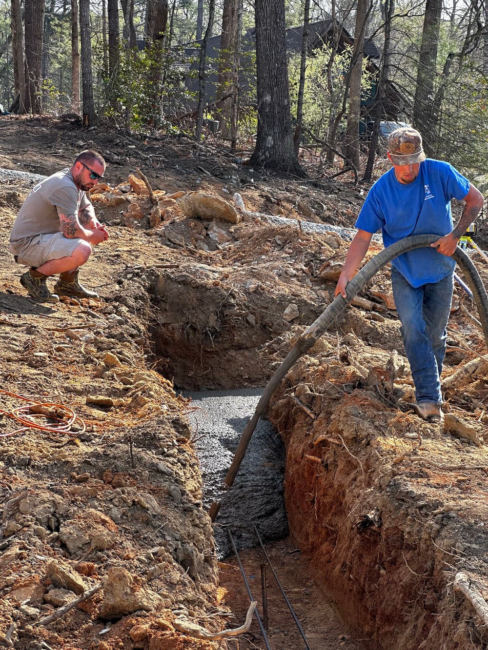 Construction workers pouring concrete in a trench in a wooded area, showcasing teamwork and labor.
