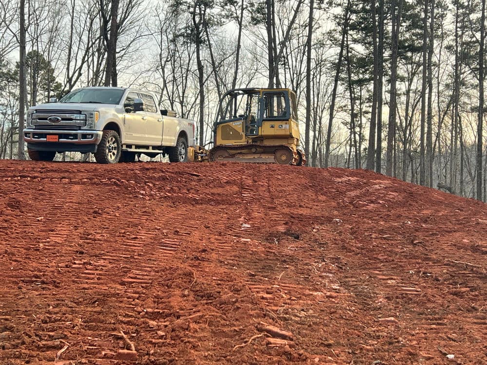 Bulldozer and pickup truck on freshly graded red dirt hill in wooded area.
