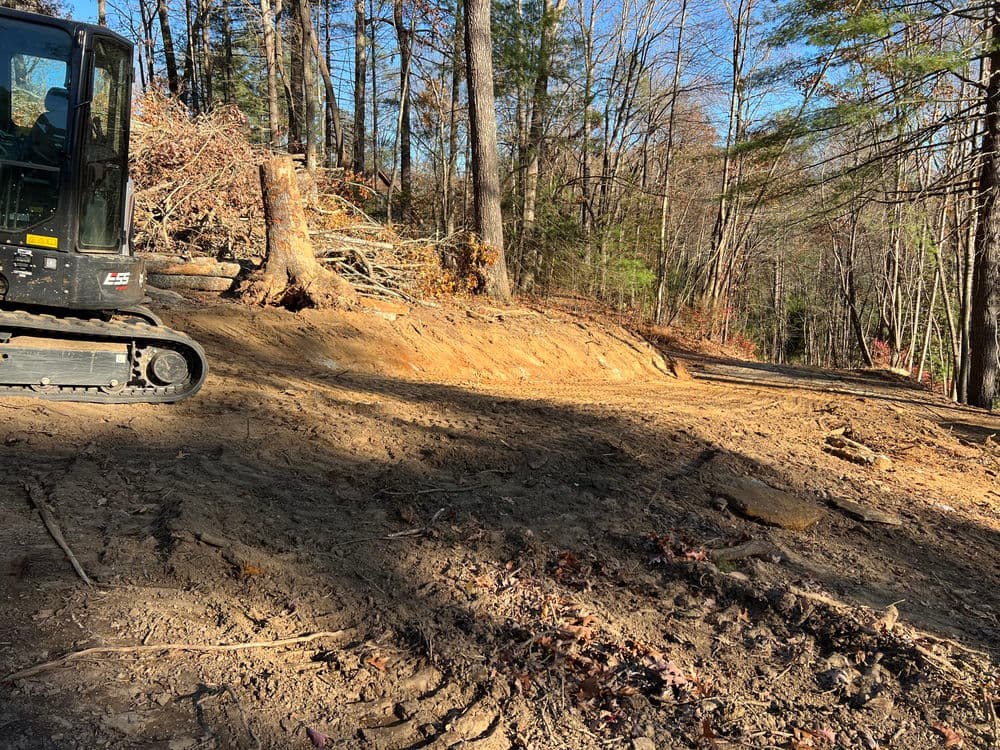 Excavator next to a cleared area in a wooded landscape, showcasing recent land development.