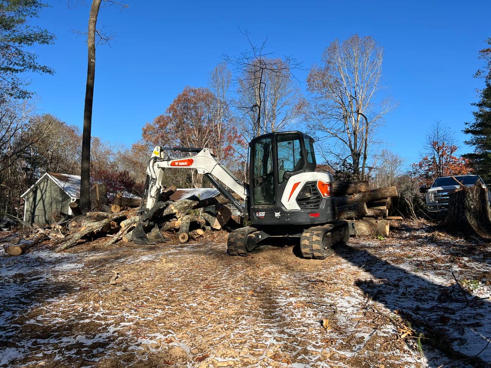Excavator clearing logs in a wooded area with autumn foliage and blue sky.