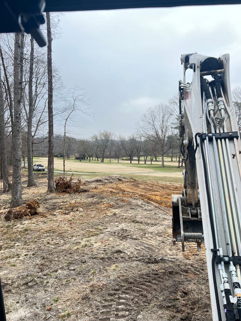 Excavator at a construction site overlooking a cleared landscape with trees and a golf course.