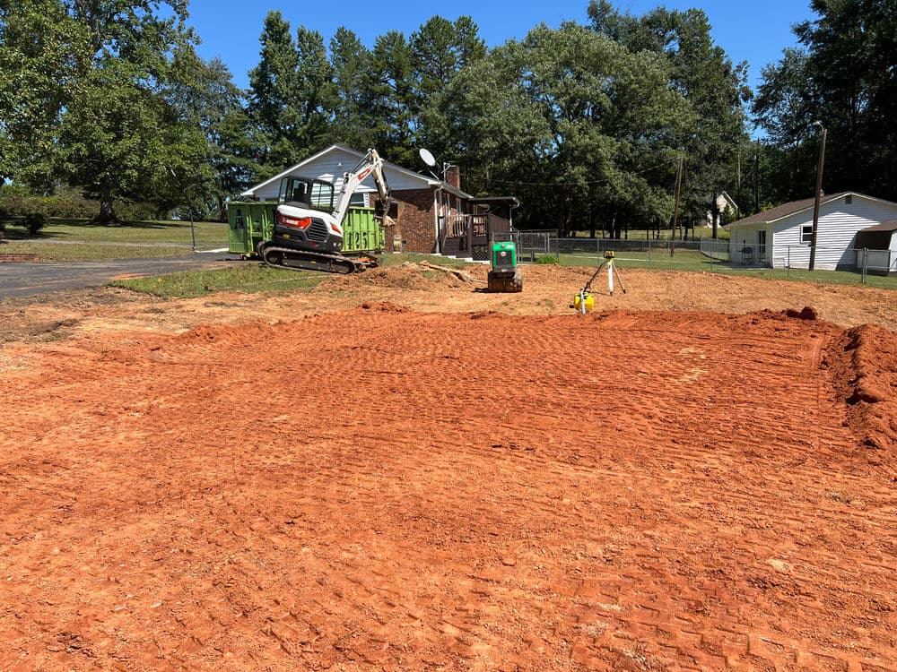 Construction site with freshly graded red dirt, machinery, and a house in the background.