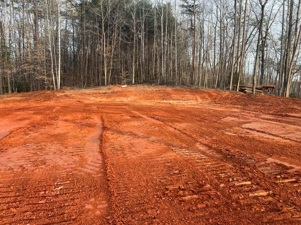 Cleared red earth with tire tracks and a forest background, indicating land preparation.