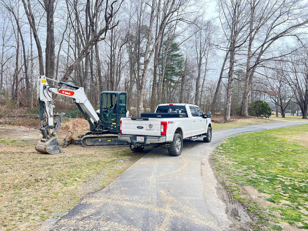 White pickup truck parked near a small excavator on a gravel path surrounded by trees.
