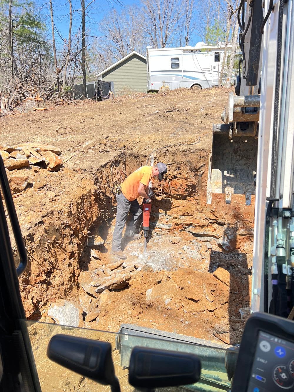 Worker operating a jackhammer in a construction site with heavy machinery and a wooded background.