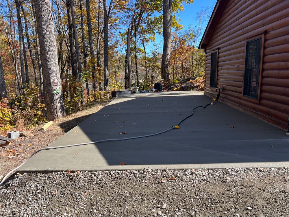 Freshly poured concrete patio behind a log cabin, surrounded by autumn trees and blue sky.