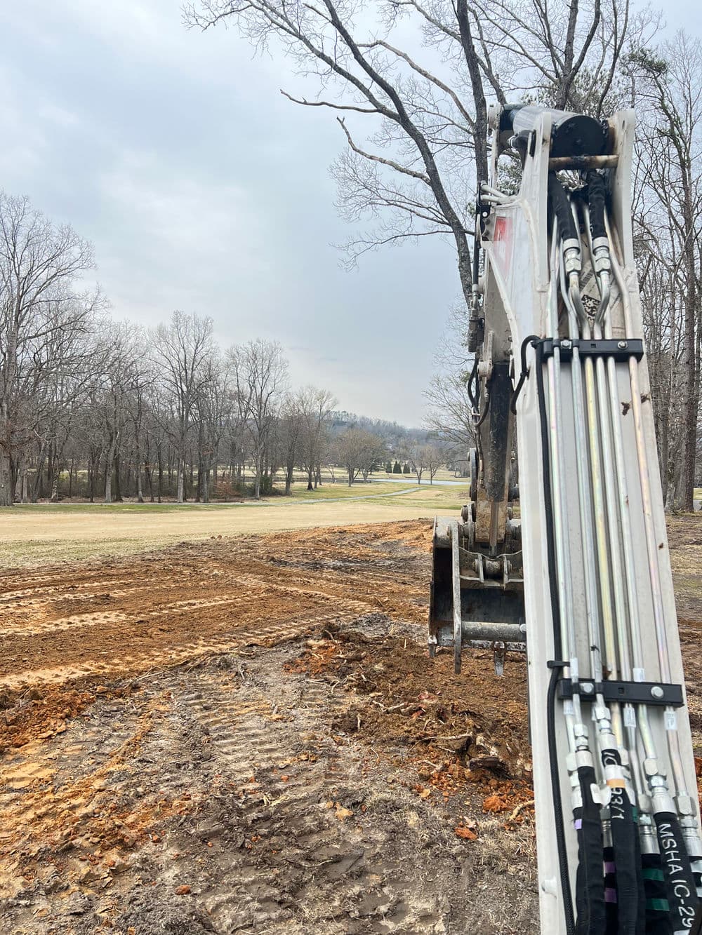 Excavator arm in a cleared area of a park, with trees and a golf course in the background.