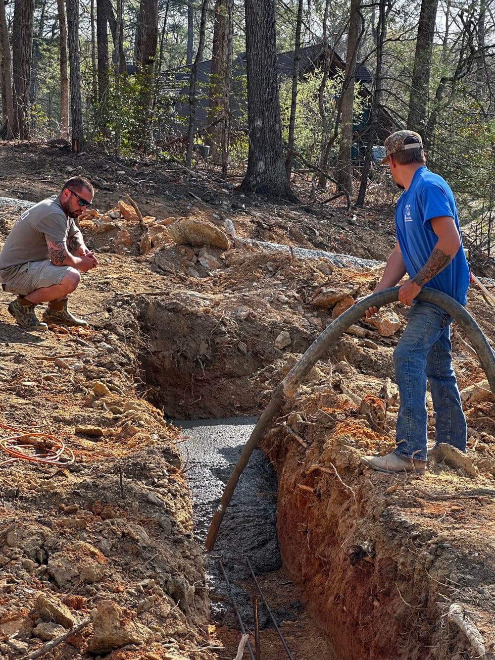 Two workers pouring concrete in a trench at a construction site surrounded by trees.