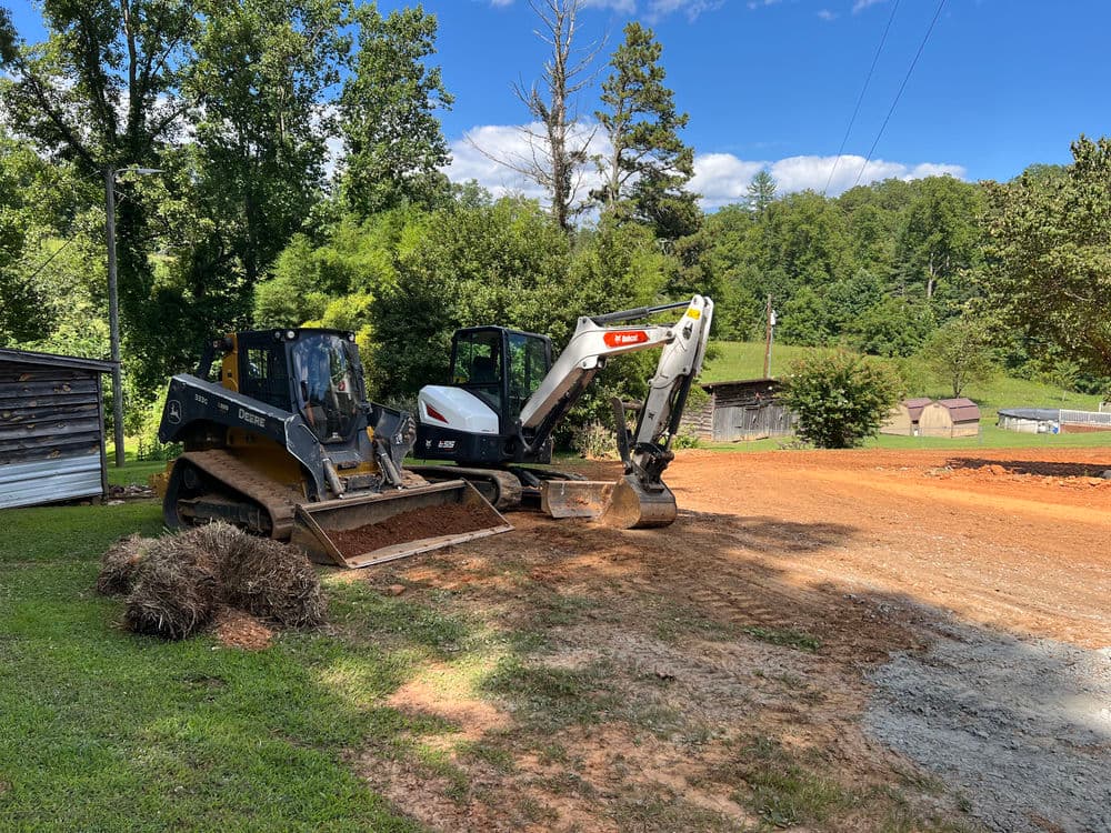 Two construction machines, a backhoe and a skid steer, working on a dirt site in a wooded area.