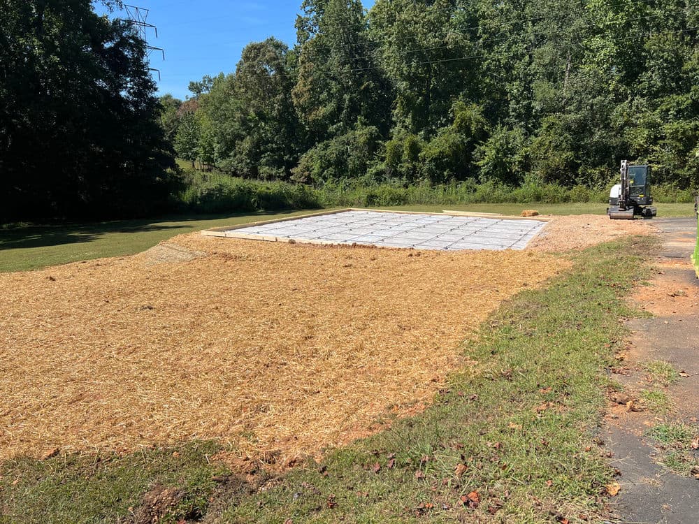 Construction site with a leveled foundation and straw mulch surrounded by trees.
