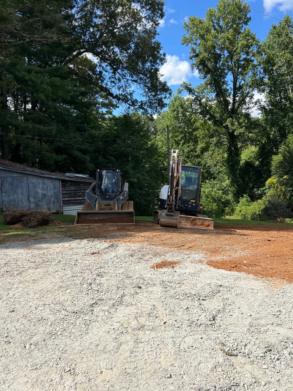 Two construction vehicles parked on a gravel area surrounded by trees and blue skies.