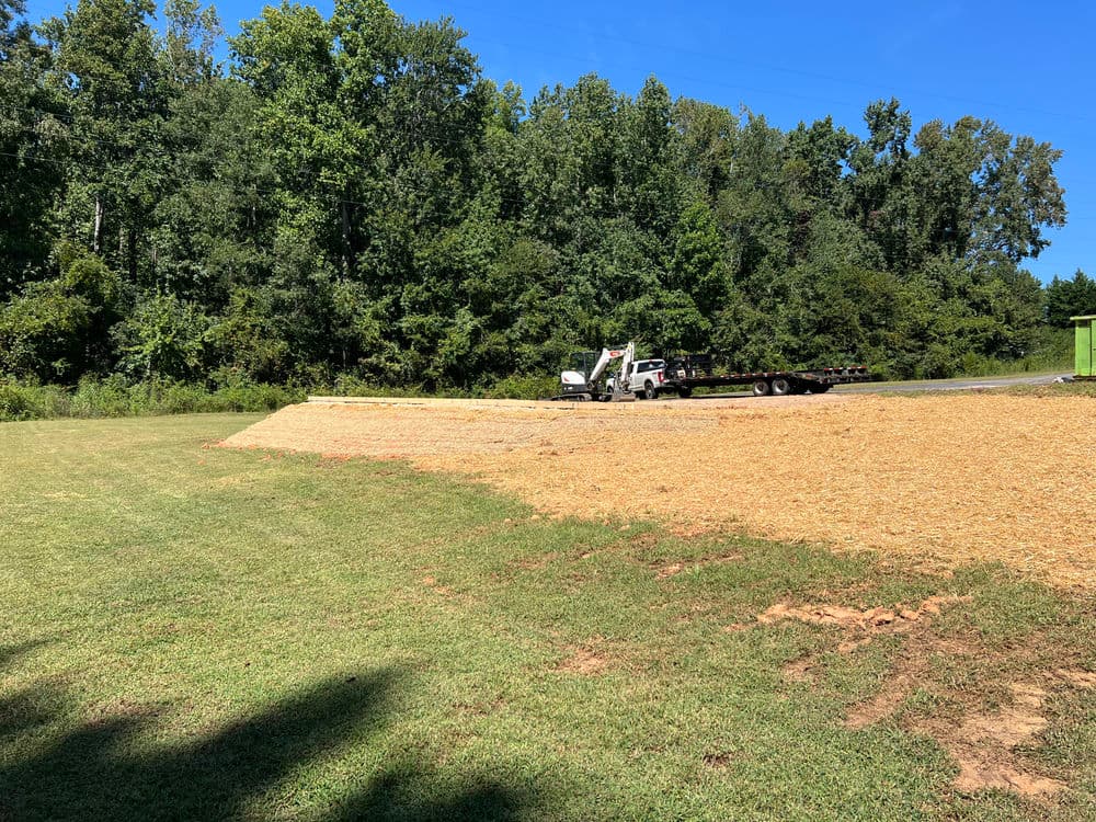 Landscape with a grassy area, gravel, and a parked truck near a wooded backdrop.
