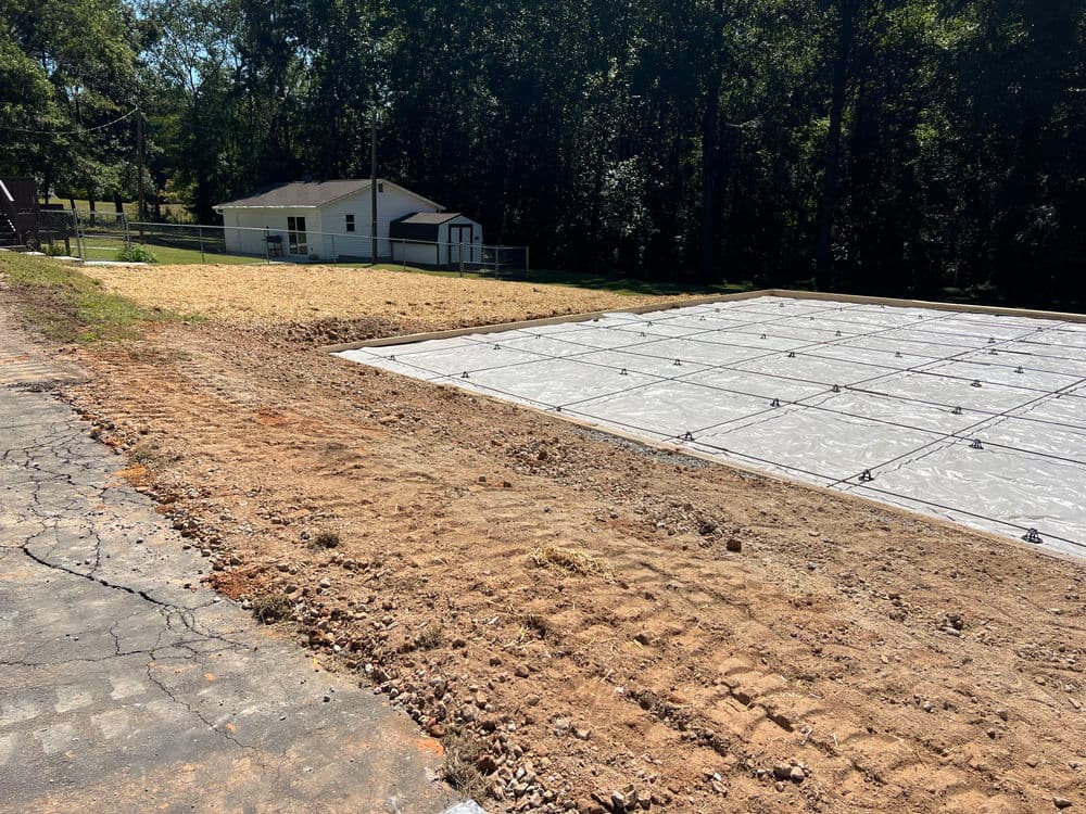 Construction site featuring a gravel area and pool cover near a white house in a wooded setting.